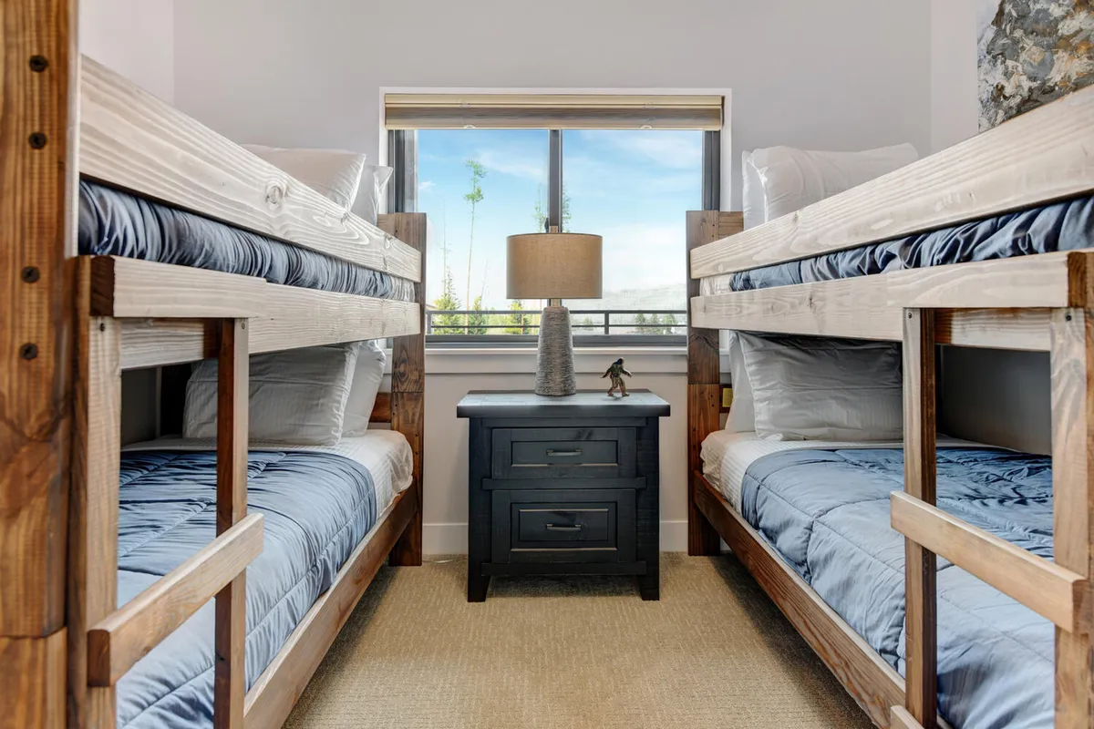 A friendly bunk room with two sets of bunk beds, a nightstand with a lamp, and a window that brings in natural light. The arrangement is practical yet playful, making it an inviting space for kids or friends.