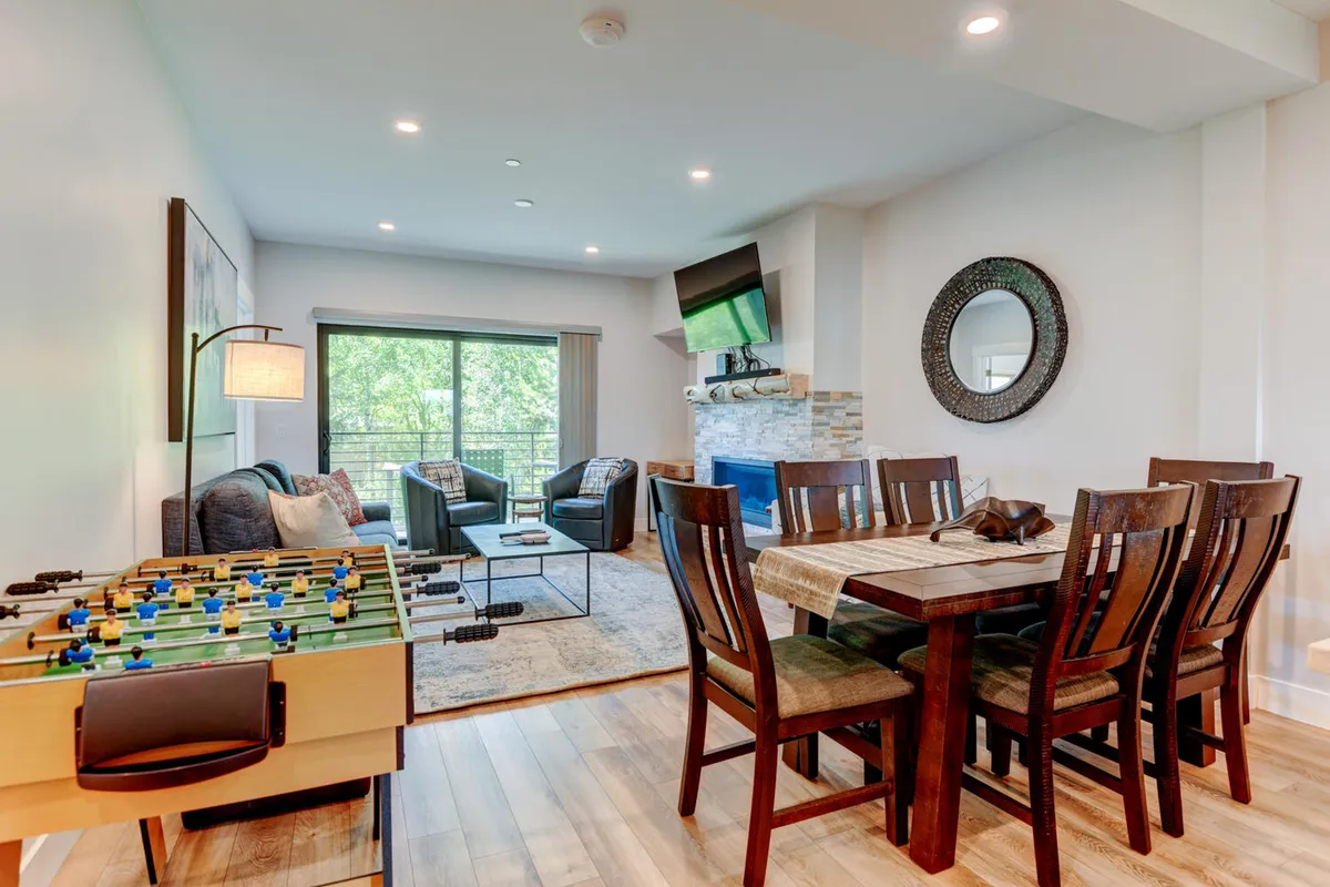 A lively open-plan living and dining area is filled with natural light and features a foosball table, spacious dining table, cozy seating area anchored by a fireplace, and a large TV. The blend of entertainment options and relaxing zones makes this space perfect for gathering with friends or family.