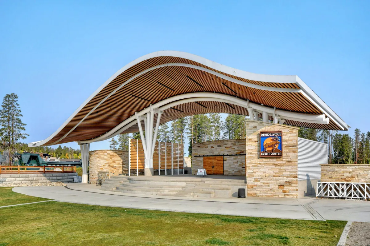 An exterior shot of a curved roof structure and stone walls, with prominent event signage. The area is set up for outdoor performances, community gatherings, and festivals, with people congregating for a live music event. The mood is energetic and communal.