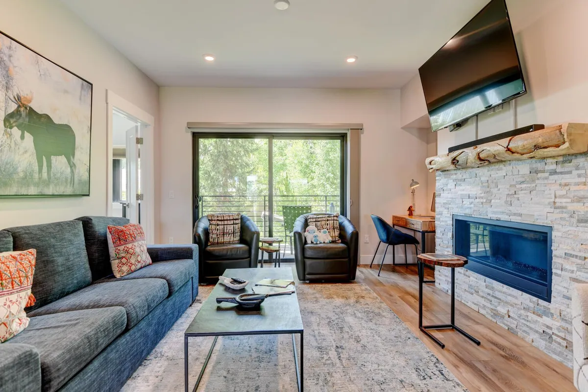 The image showcases a stylish, inviting living room featuring a plush sofa with decorative pillows, a glowing fireplace, expansive windows offering views of the outdoors, and a striking moose artwork. The scene radiates warmth and comfort, inviting guests to relax, socialize, and enjoy the cozy mountain ambiance.