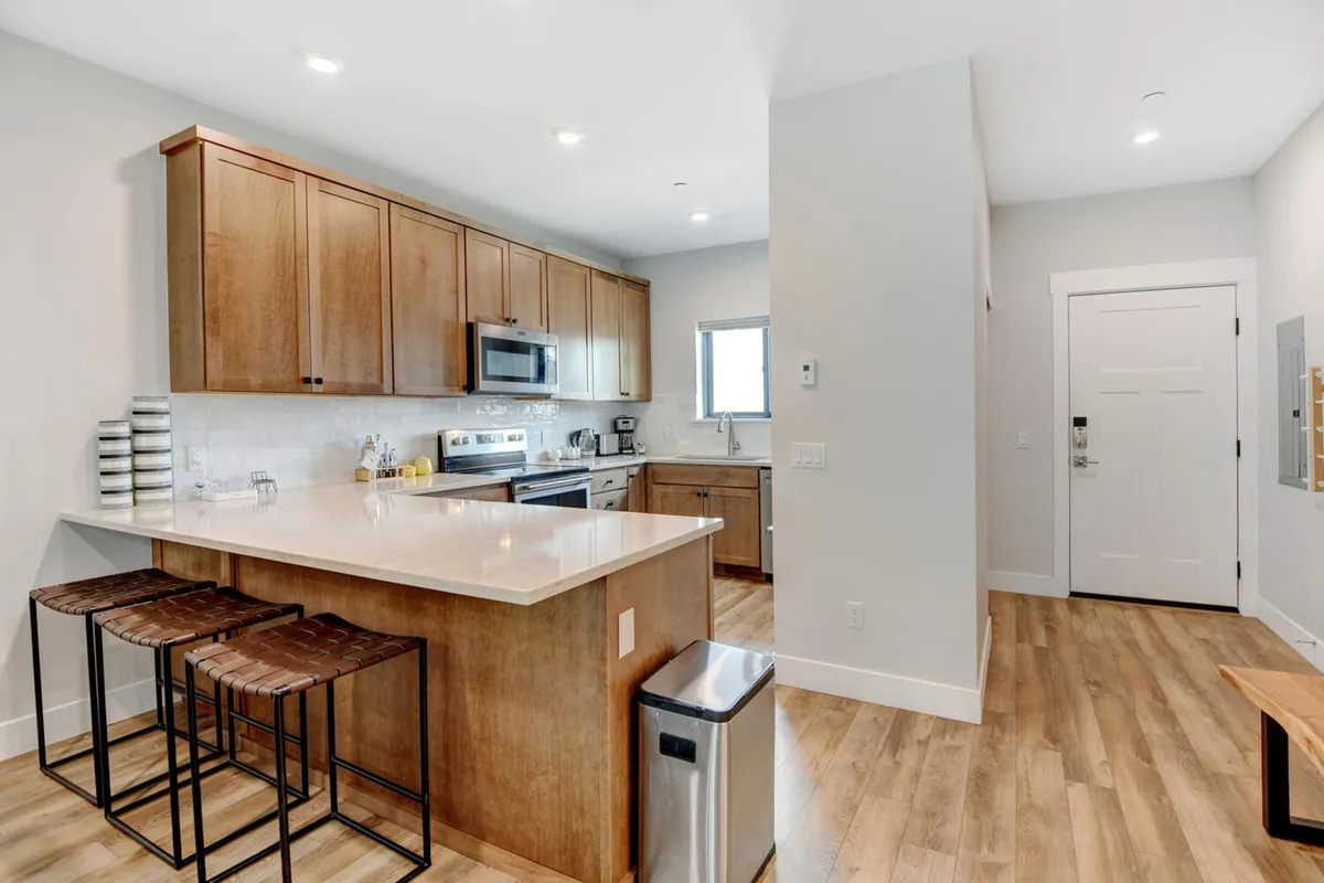 A modern kitchen area showcases a kitchen island with bar stools, sleek stainless steel appliances, wooden cabinetry, and a large window letting in mountain light. The space feels welcoming and functional, ideal for family meals or group gatherings.