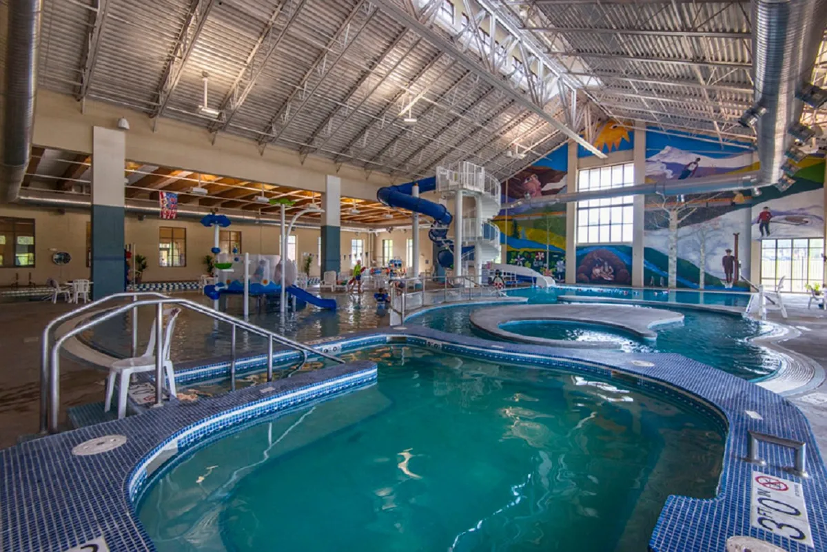 A vibrant indoor aquatic center bustling with activity. The image shows colorful water slides winding above sparkling swimming pools, with children playing and families relaxing poolside. A lively mural decorates the background, enhancing the fun, resort-like atmosphere.