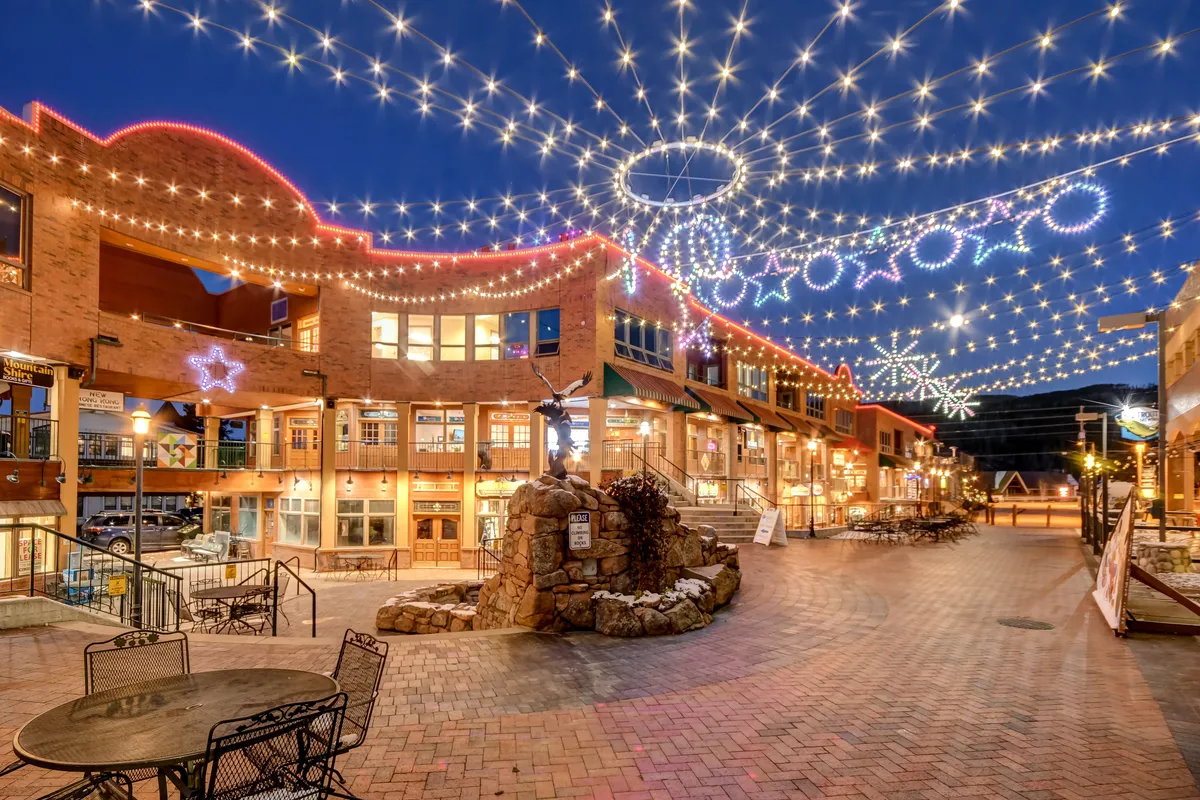 A bustling downtown street scene featuring decorative lights strung overhead, a central fountain as a focal point, and a lively array of storefronts lining the walkway. People can be seen dining, shopping, and strolling, creating a vibrant community atmosphere.