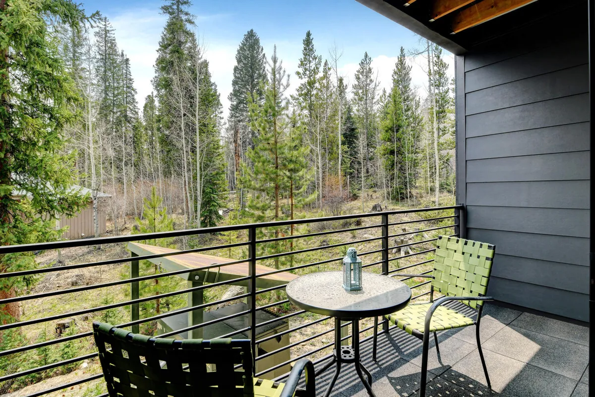 A peaceful private deck is surrounded by mature trees and features a small round table with two green chairs, providing an inviting spot for coffee, conversation, or simply soaking in the Colorado outdoors.