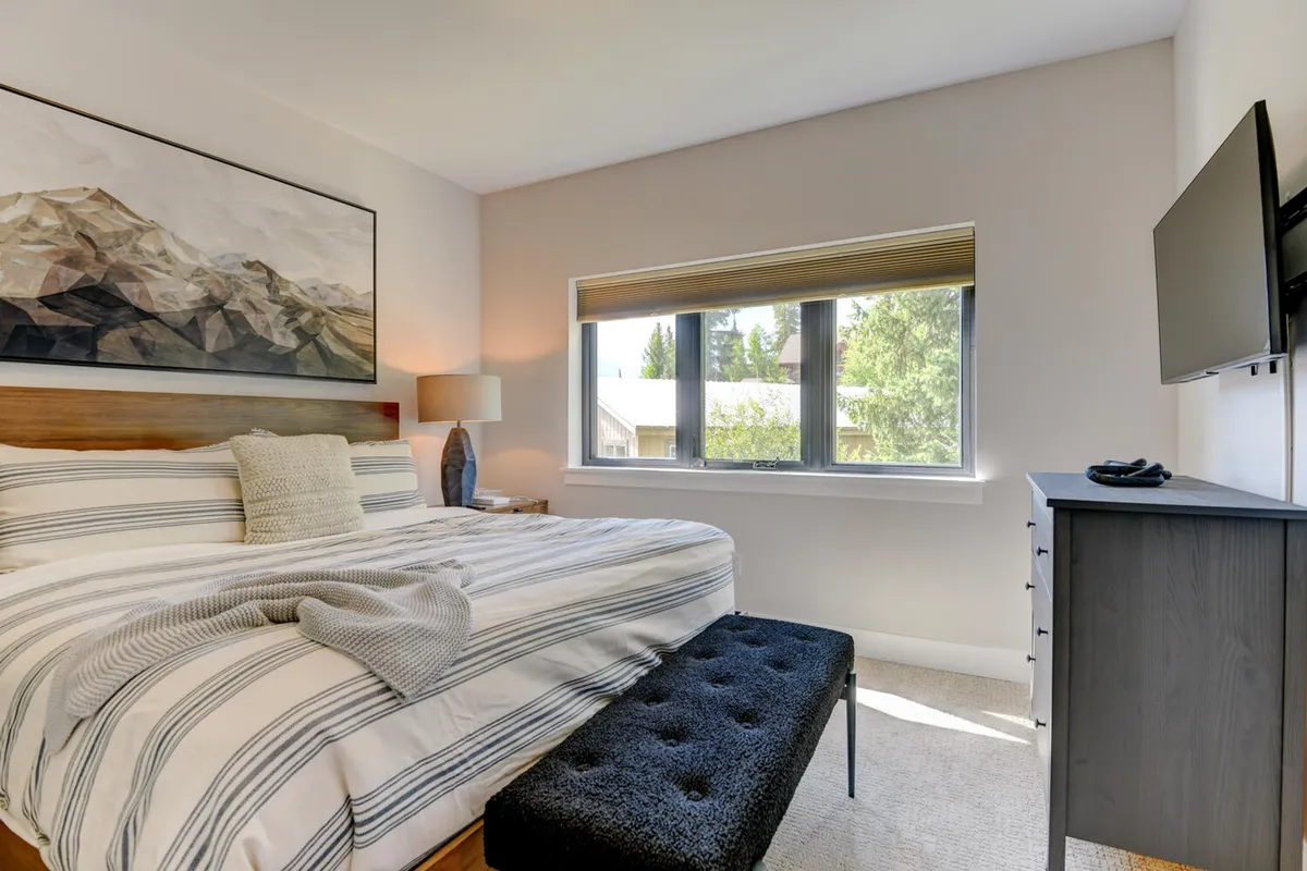 A serene bedroom with striped bedding, a large mountain artwork above the bed, a window view letting in natural light, and a cozy bench—offering a restful retreat after group adventures.