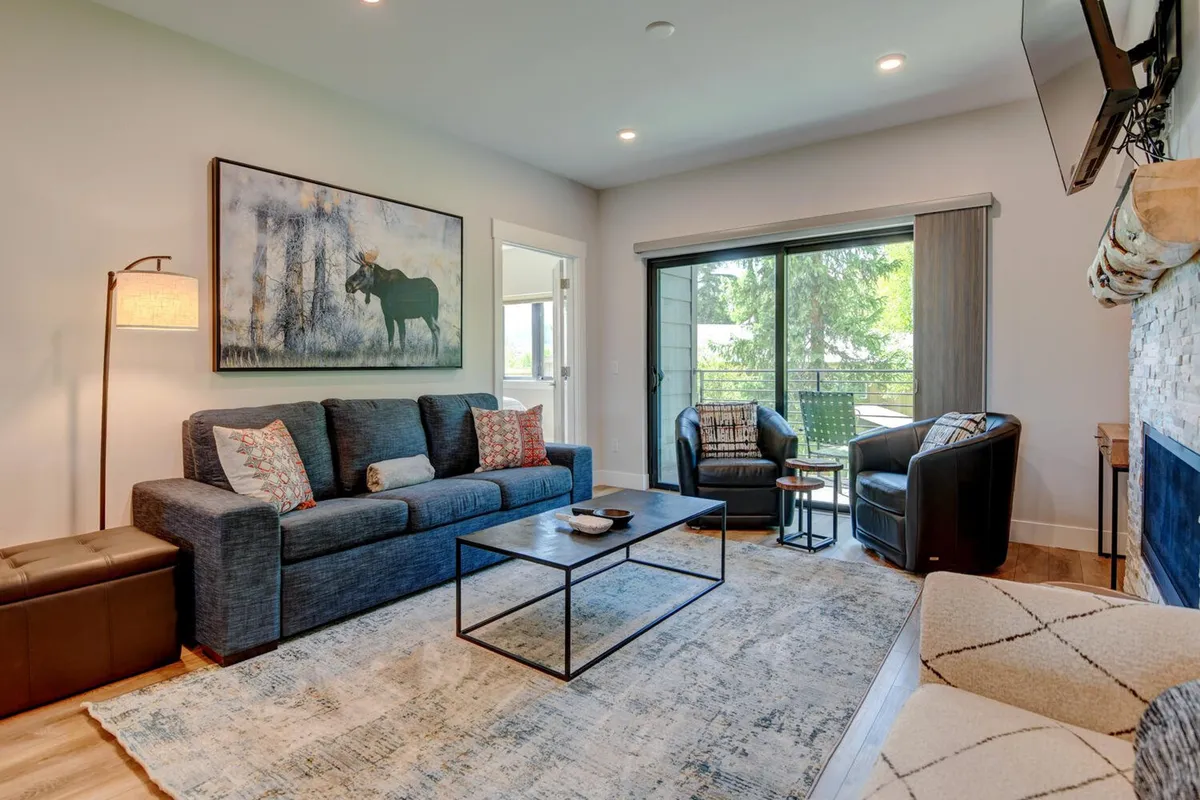A cozy living room with a sofa, coffee table, window showcasing the outdoors, a fireplace, and artwork. The large window draws attention to the scenery outside, reinforcing a connection to the changing seasons and activities beyond the home.