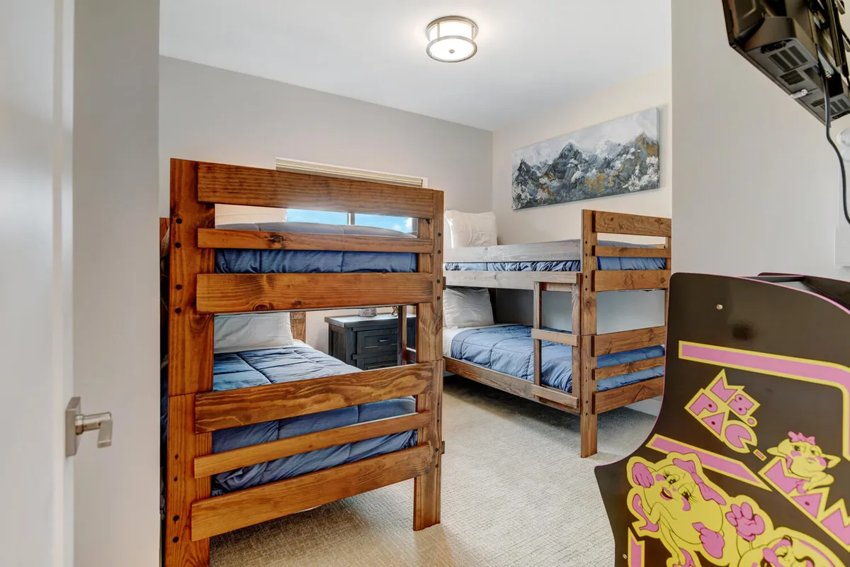 A lively bunk room featuring cozy bunk beds, playful artwork, a nightstand, and a classic arcade machine. The space radiates fun and is designed to encourage both restful sleep and interactive gaming, appealing to children and teens alike.