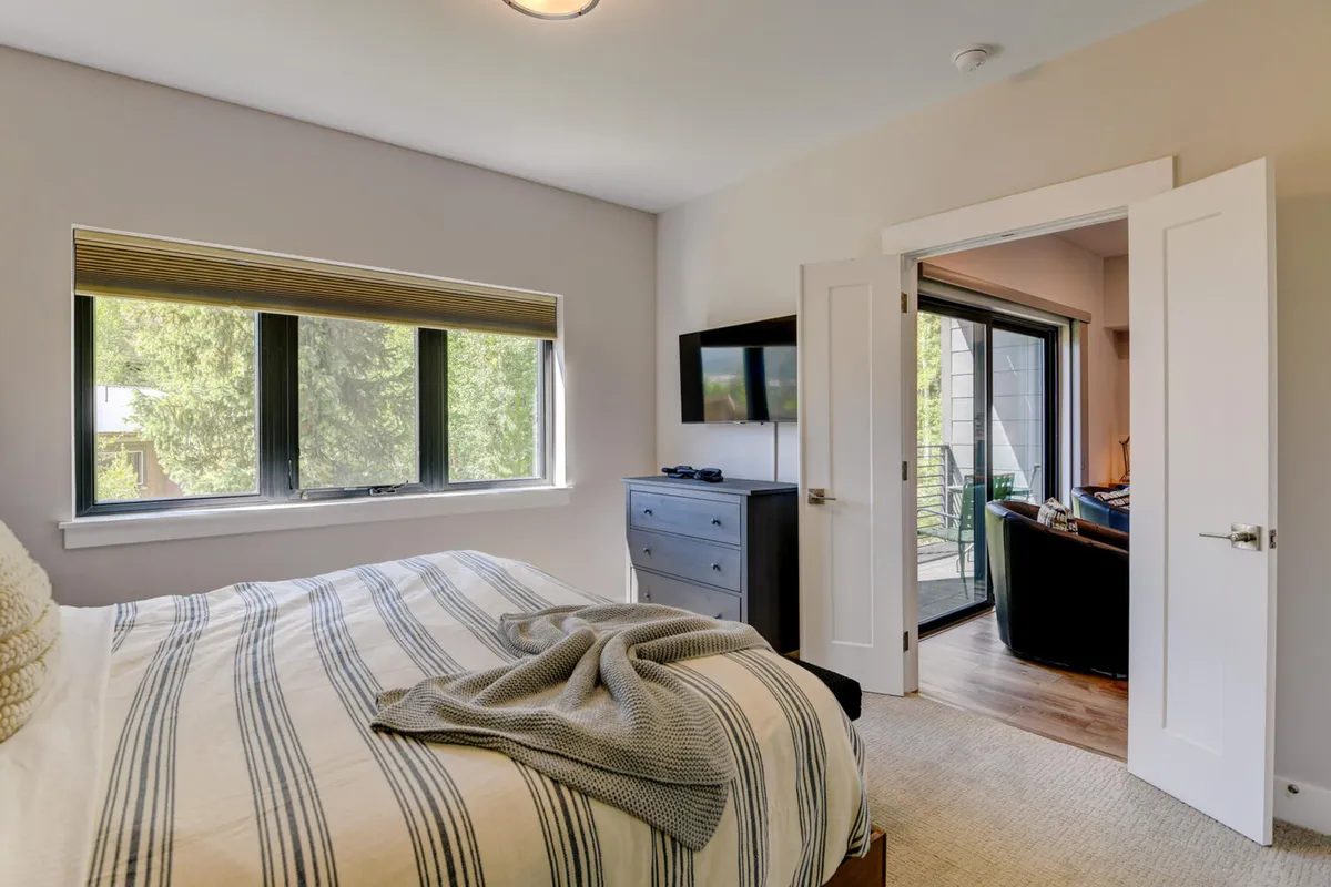 A serene bedroom with a bed featuring striped bedding, large windows letting in natural light, a television opposite the bed, and a door leading outside. The peaceful decor is accented with soft tones and cozy textures.